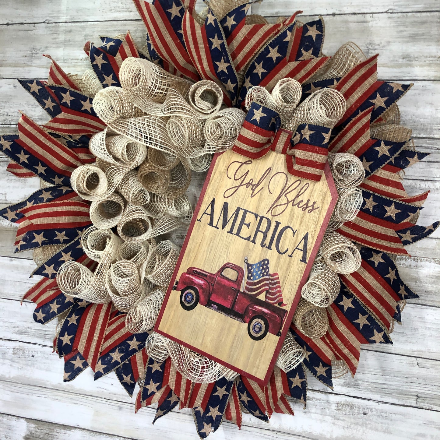 God Bless America! This farmhouse style wreath has a beige base and a very simple ribbon design. The American Flag accent ribbon has a rustic and worn look. The large sign features an old style pickup truck flying the American Flag out of the back with the words, "God Bless America" The details of how everything comes together makes this the perfect patriotic farmhouse wreath.