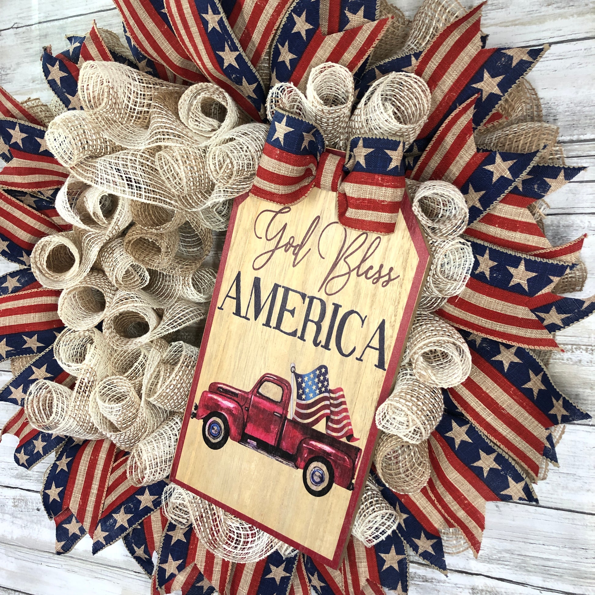 God Bless America! This farmhouse style wreath has a beige base and a very simple ribbon design. The American Flag accent ribbon has a rustic and worn look. The large sign features an old style pickup truck flying the American Flag out of the back with the words, "God Bless America" The details of how everything comes together makes this the perfect patriotic farmhouse wreath.