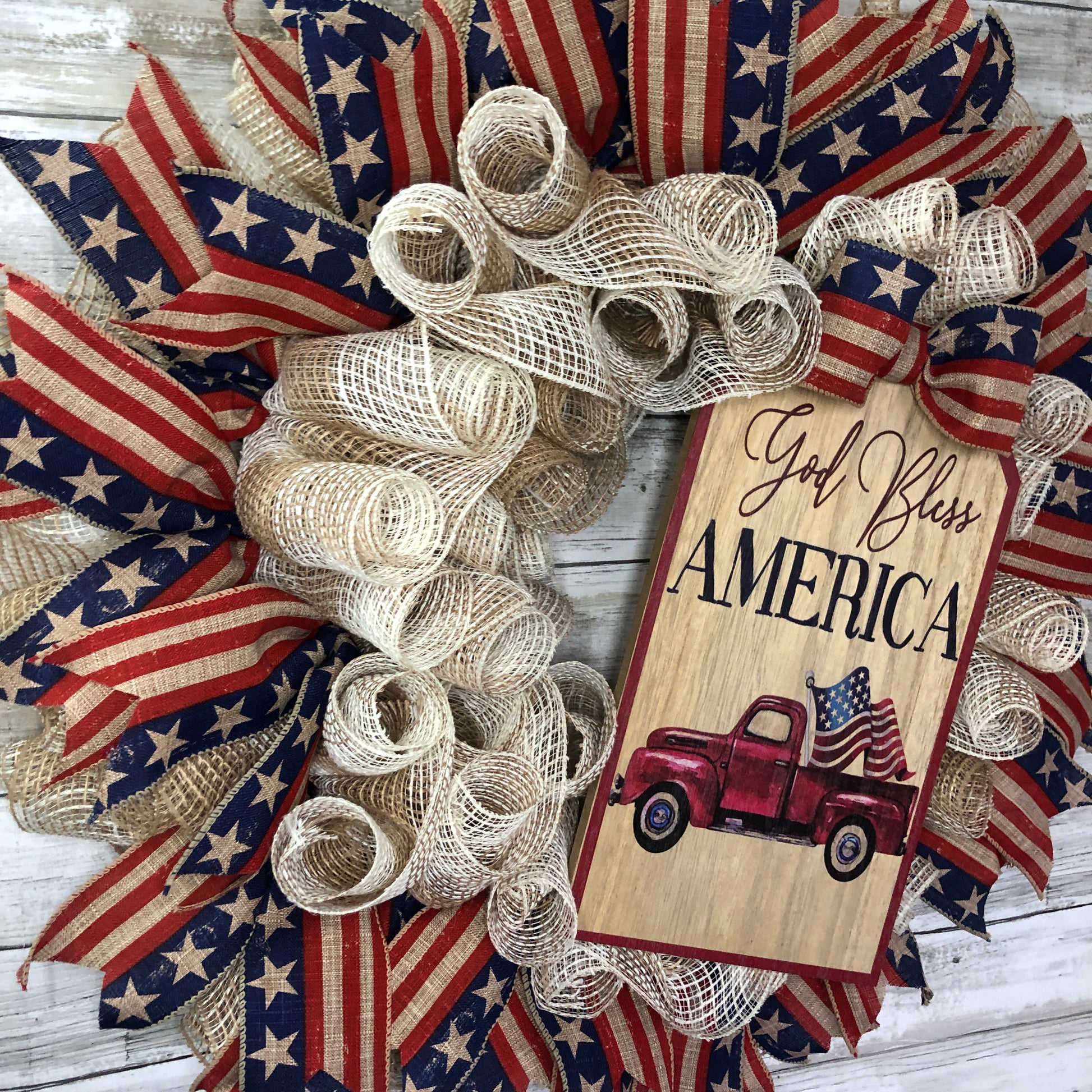 God Bless America! This farmhouse style wreath has a beige base and a very simple ribbon design. The American Flag accent ribbon has a rustic and worn look. The large sign features an old style pickup truck flying the American Flag out of the back with the words, "God Bless America" The details of how everything comes together makes this the perfect patriotic farmhouse wreath.