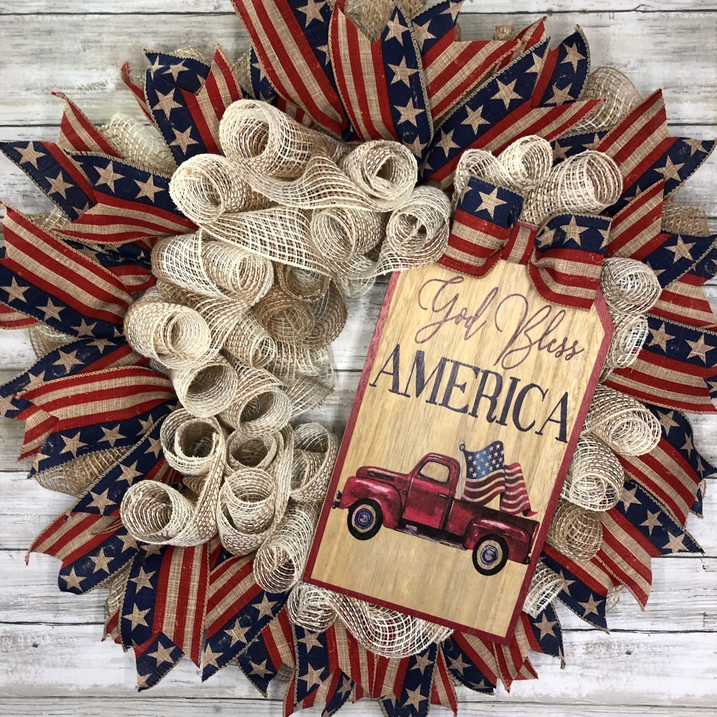 God Bless America! This farmhouse style wreath has a beige base and a very simple ribbon design. The American Flag accent ribbon has a rustic and worn look. The large sign features an old style pickup truck flying the American Flag out of the back with the words, "God Bless America" The details of how everything comes together makes this the perfect patriotic farmhouse wreath.