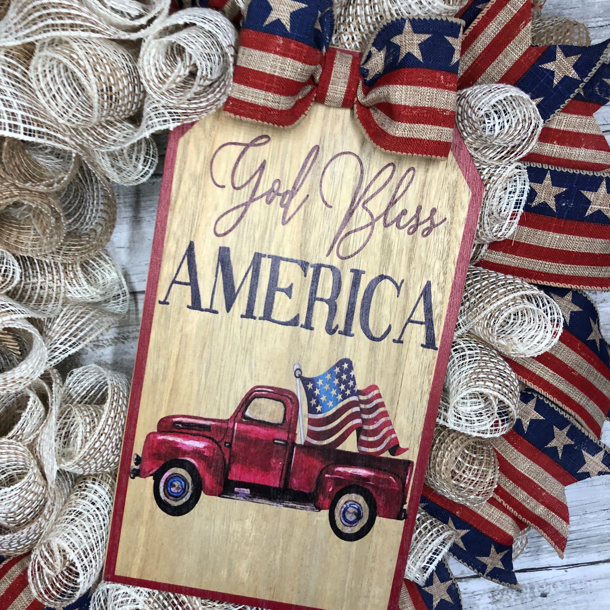 God Bless America! This farmhouse style wreath has a beige base and a very simple ribbon design. The American Flag accent ribbon has a rustic and worn look. The large sign features an old style pickup truck flying the American Flag out of the back with the words, "God Bless America" The details of how everything comes together makes this the perfect patriotic farmhouse wreath.