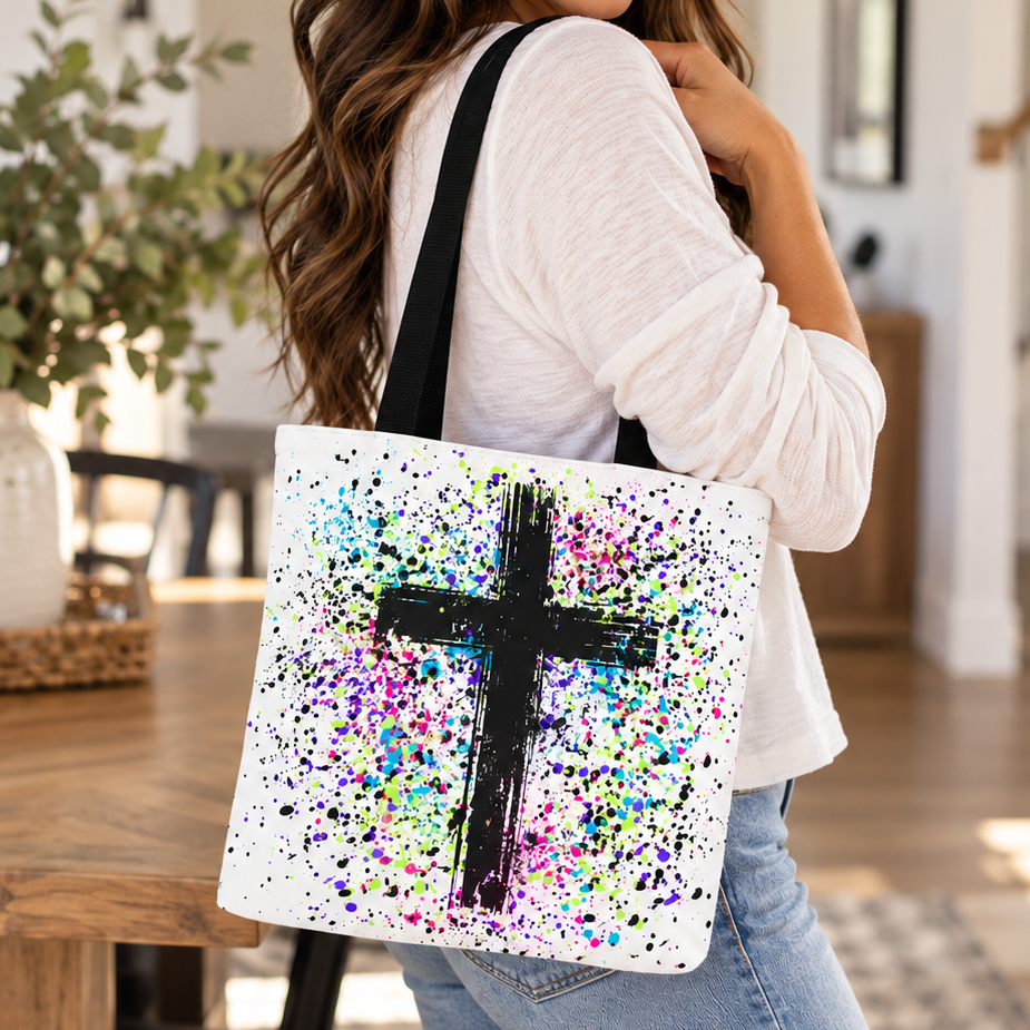 A woman wearing a white long-sleeve top and light blue jeans carries a white canvas tote bag with a black brush-stroke cross and a colorful multicolor paint splatter design on the front, set in a bright, modern home interior.